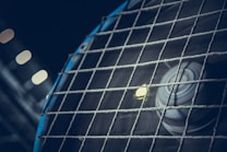 A close-up view of a spinning industrial fan, partially covered by a wire mesh fence. The fan blades appear slightly blurred due to their motion. The lighting creates a moody atmosphere with a few out-of-focus lights in the background.