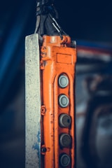 An industrial control pendant with an orange casing and multiple grey metallic buttons arranged vertically. The control pendant is attached to a metal structure with visible scratches and wear. The background is blurred, emphasizing the focus on the industrial equipment.