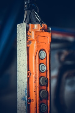 An industrial control pendant with an orange casing and multiple grey metallic buttons arranged vertically. The control pendant is attached to a metal structure with visible scratches and wear. The background is blurred, emphasizing the focus on the industrial equipment.