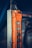 An industrial control pendant with an orange casing and multiple grey metallic buttons arranged vertically. The control pendant is attached to a metal structure with visible scratches and wear. The background is blurred, emphasizing the focus on the industrial equipment.