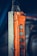 An industrial control pendant with an orange casing and multiple grey metallic buttons arranged vertically. The control pendant is attached to a metal structure with visible scratches and wear. The background is blurred, emphasizing the focus on the industrial equipment.