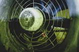 A close-up view of an industrial fan with a metal grille, showing visible signs of dirt and wear. The fan blades are behind the grille, and the motor housing is prominently featured in the center.