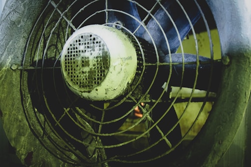 A close-up view of an industrial fan with a metal grille, showing visible signs of dirt and wear. The fan blades are behind the grille, and the motor housing is prominently featured in the center.