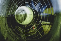 A close-up view of an industrial fan with a metal grille, showing visible signs of dirt and wear. The fan blades are behind the grille, and the motor housing is prominently featured in the center.