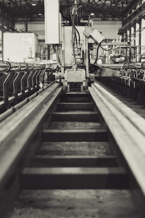 A monochrome industrial scene featuring a large piece of machinery in the center, flanked by symmetrical metal tracks on either side. The background reveals a spacious warehouse with structural beams and additional equipment. The setting appears to be a manufacturing or processing facility.