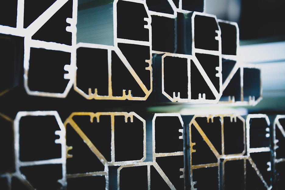 Several aluminum profiles are stacked together, showcasing their hollow, geometric shapes with precise, angular lines and smooth surfaces.