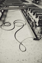 A monochrome image of an industrial setting, featuring long metal rollers and a concrete floor with a coiled cable. The machinery suggests a factory or manufacturing environment, and the cable lies casually, adding a sense of untidiness.