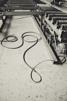 A monochrome image of an industrial setting, featuring long metal rollers and a concrete floor with a coiled cable. The machinery suggests a factory or manufacturing environment, and the cable lies casually, adding a sense of untidiness.
