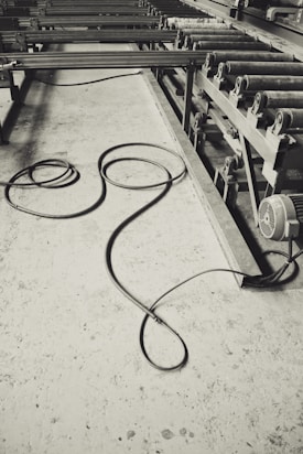 A monochrome image of an industrial setting, featuring long metal rollers and a concrete floor with a coiled cable. The machinery suggests a factory or manufacturing environment, and the cable lies casually, adding a sense of untidiness.