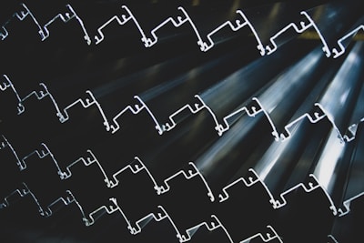 Rows of metallic extrusions with intricate shapes, stacked together to form a textured and rhythmic pattern. The lighting highlights the contours and edges, creating a sense of depth and complexity.