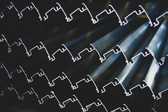 Rows of metallic extrusions with intricate shapes, stacked together to form a textured and rhythmic pattern. The lighting highlights the contours and edges, creating a sense of depth and complexity.
