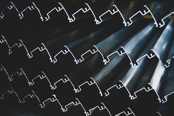 Rows of metallic extrusions with intricate shapes, stacked together to form a textured and rhythmic pattern. The lighting highlights the contours and edges, creating a sense of depth and complexity.