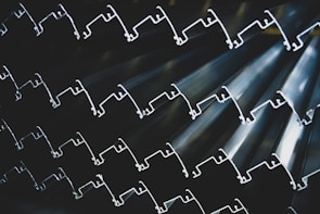 Rows of metallic extrusions with intricate shapes, stacked together to form a textured and rhythmic pattern. The lighting highlights the contours and edges, creating a sense of depth and complexity.