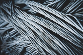 A close-up view of a dense bundle of twisted, metallic wires, exhibiting a slightly weathered texture with hints of rust. The wires intersect and overlap in a chaotic yet organized manner, creating a sense of depth and complexity.
