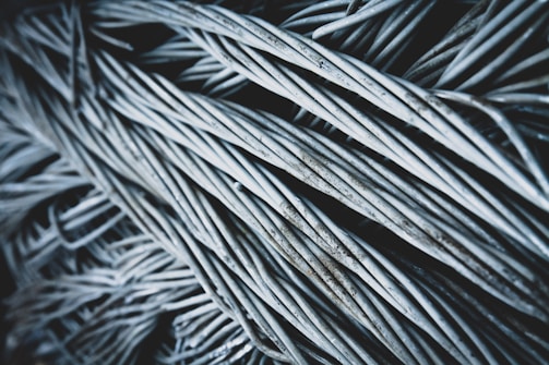 A close-up view of a dense bundle of twisted, metallic wires, exhibiting a slightly weathered texture with hints of rust. The wires intersect and overlap in a chaotic yet organized manner, creating a sense of depth and complexity.