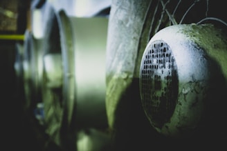 Close-up view of a weathered and slightly rusted industrial fan or ventilation system. The focus is on the front protective grill with blurred background elements featuring similar equipment. The lighting is dim, providing a sense of age and usage.