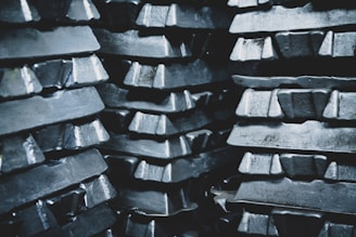 Stacks of metallic ingots, arranged in several layers, appearing shiny and rough in texture. The ingots are tightly packed, creating a pattern of repeating shapes and reflections.