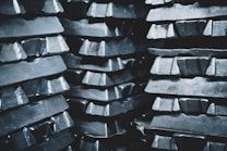 Stacks of metallic ingots, arranged in several layers, appearing shiny and rough in texture. The ingots are tightly packed, creating a pattern of repeating shapes and reflections.