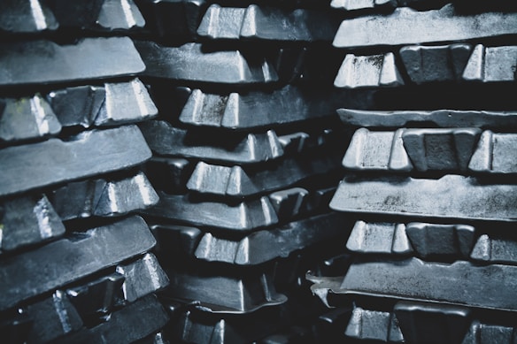 Stacks of metallic ingots, arranged in several layers, appearing shiny and rough in texture. The ingots are tightly packed, creating a pattern of repeating shapes and reflections.