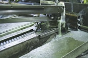 A metal-cutting machine in action, with a saw blade slicing through pipes and coolant fluid being applied to reduce heat and friction. The environment has an industrial and mechanical feel with visible metal shavings and gears.