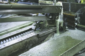 A metal-cutting machine in action, with a saw blade slicing through pipes and coolant fluid being applied to reduce heat and friction. The environment has an industrial and mechanical feel with visible metal shavings and gears.