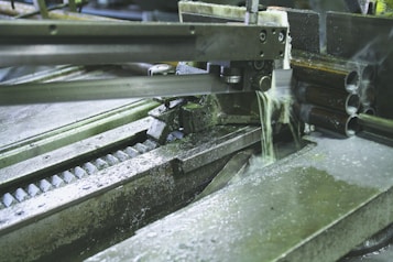 A metal-cutting machine in action, with a saw blade slicing through pipes and coolant fluid being applied to reduce heat and friction. The environment has an industrial and mechanical feel with visible metal shavings and gears.