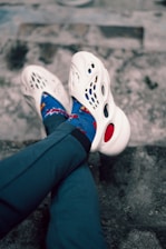 A pair of legs crossed at the ankles, wearing white foam shoes with multiple cutouts and colorful blue socks featuring a playful design. The legs are clad in dark blue or black pants. The background consists of a textured stone or concrete surface.