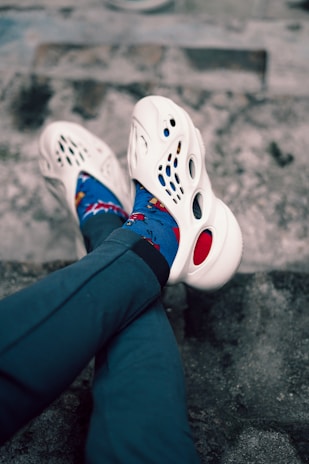 A pair of legs crossed at the ankles, wearing white foam shoes with multiple cutouts and colorful blue socks featuring a playful design. The legs are clad in dark blue or black pants. The background consists of a textured stone or concrete surface.