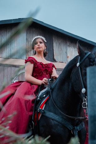 A woman in an elegant red dress sits confidently atop a black horse, positioned in front of a rustic, weathered building. She wears a decorative headpiece and long earrings, exuding a regal presence.