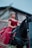A woman in an elegant red dress sits confidently atop a black horse, positioned in front of a rustic, weathered building. She wears a decorative headpiece and long earrings, exuding a regal presence.