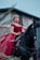 A woman in an elegant red dress sits confidently atop a black horse, positioned in front of a rustic, weathered building. She wears a decorative headpiece and long earrings, exuding a regal presence.