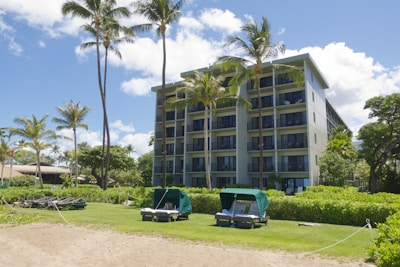 A multi-story resort building surrounded by palm trees is set against a backdrop of a clear blue sky with scattered clouds. In front of the building, there is a grassy area with lounge chairs and cabanas, bordered by a sandy path. The scene suggests a tropical, vacation-like setting.
