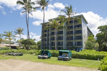 A multi-story resort building surrounded by palm trees is set against a backdrop of a clear blue sky with scattered clouds. In front of the building, there is a grassy area with lounge chairs and cabanas, bordered by a sandy path. The scene suggests a tropical, vacation-like setting.