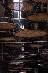 Circular bamboo trays are suspended in a narrow, dimly lit alleyway. The trays are used for drying food, and a light rain is visible, with droplets falling against the dark background. Metal rods support the trays, and wires help hold them in place.