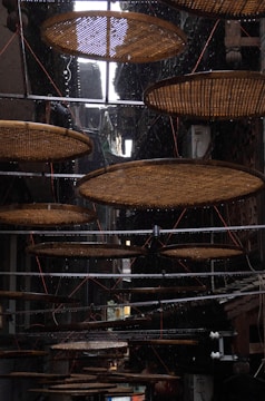 Circular bamboo trays are suspended in a narrow, dimly lit alleyway. The trays are used for drying food, and a light rain is visible, with droplets falling against the dark background. Metal rods support the trays, and wires help hold them in place.
