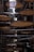Circular bamboo trays are suspended in a narrow, dimly lit alleyway. The trays are used for drying food, and a light rain is visible, with droplets falling against the dark background. Metal rods support the trays, and wires help hold them in place.