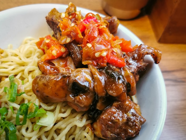 A serving of noodles topped with savory, glazed pieces of meat and garnished with vibrant red chili peppers and sliced green onions.