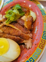 Sliced roasted duck is placed on a decorative red plate with a colorful border, accompanied by halves of a boiled egg and garnished with fresh cilantro leaves.