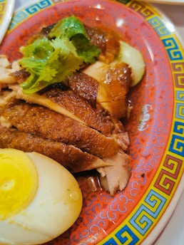 Sliced roasted duck is placed on a decorative red plate with a colorful border, accompanied by halves of a boiled egg and garnished with fresh cilantro leaves.