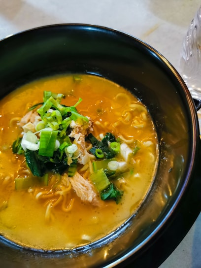 A bowl of ramen soup garnished with fresh green onions and leafy greens. The broth is rich and golden, with a few pieces of tender meat visible on top. The noodles are submerged beneath the surface, adding to the aromatic composition.