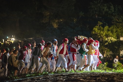 A group of people dressed in colorful, traditional attire walk together at night. They appear to be participating in a procession, with some carrying an object on their shoulders covered in a red and gold cloth. The setting is outdoors, surrounded by greenery, with lights visible in the background.