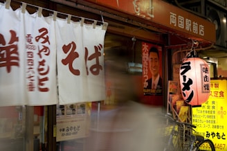 A Japanese storefront features white curtains with red kanji characters, a red and white lantern with black kanji characters, and a blurred figure walking past. A bicycle is parked nearby, and various posters and menus are visible, including a poster with a man in a suit.