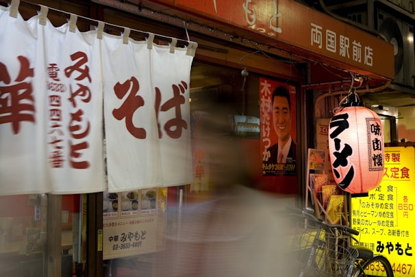 A Japanese storefront features white curtains with red kanji characters, a red and white lantern with black kanji characters, and a blurred figure walking past. A bicycle is parked nearby, and various posters and menus are visible, including a poster with a man in a suit.