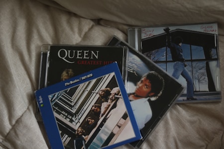 A collection of music CDs is placed on a quilted fabric surface. The CDs feature iconic artists and albums, including Queen's Greatest Hits, a Billy Joel album titled Glass Houses, a Michael Jackson album, and The Beatles' 1967-1970 compilation. The covers display a range of album artwork, from band photos to portraits of individual artists.