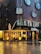 A modern hotel building with circular windows arranged in a grid pattern on its dark, glossy facade. The ground floor features large glass windows showcasing a lobby interior with warm decorative lights. The wet street reflects the building, hinting at recent rainfall. A street sign and umbrella add to the urban scene.