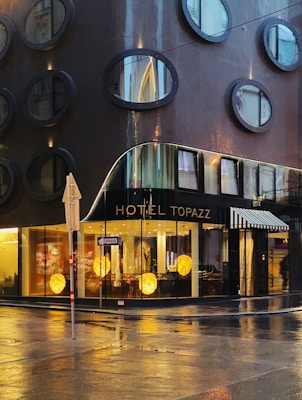 A modern hotel building with circular windows arranged in a grid pattern on its dark, glossy facade. The ground floor features large glass windows showcasing a lobby interior with warm decorative lights. The wet street reflects the building, hinting at recent rainfall. A street sign and umbrella add to the urban scene.