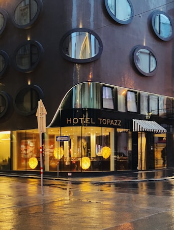 A modern hotel building with circular windows arranged in a grid pattern on its dark, glossy facade. The ground floor features large glass windows showcasing a lobby interior with warm decorative lights. The wet street reflects the building, hinting at recent rainfall. A street sign and umbrella add to the urban scene.