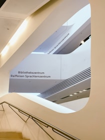 An architectural interior with curved white walls and clean lines, featuring a staircase with a metal handrail. The text 'Bibliothekszentrum' and 'Raiffeisen Sprachlernzentrum' is visible on the walls, suggesting a library or language center. The design is modern and minimalistic, emphasizing open space and light.