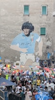 A large mural depicting a soccer player in a blue jersey is painted on a tall building. Below the mural, a bustling street market is set up with people shopping among numerous stalls. Brightly colored flags are strung across the scene, adding vibrance. Several people are browsing or purchasing merchandise, including sports jerseys.
