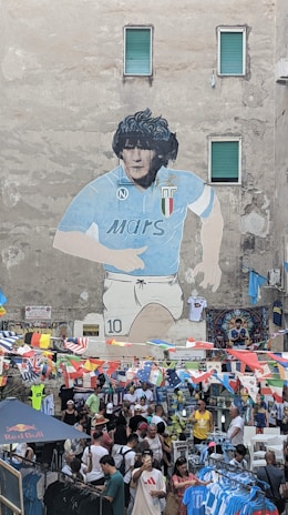 A large mural depicting a soccer player in a blue jersey is painted on a tall building. Below the mural, a bustling street market is set up with people shopping among numerous stalls. Brightly colored flags are strung across the scene, adding vibrance. Several people are browsing or purchasing merchandise, including sports jerseys.